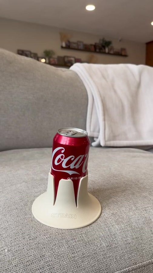 A full can standing in a white Steadi on a couch cushion, a living room behind it.